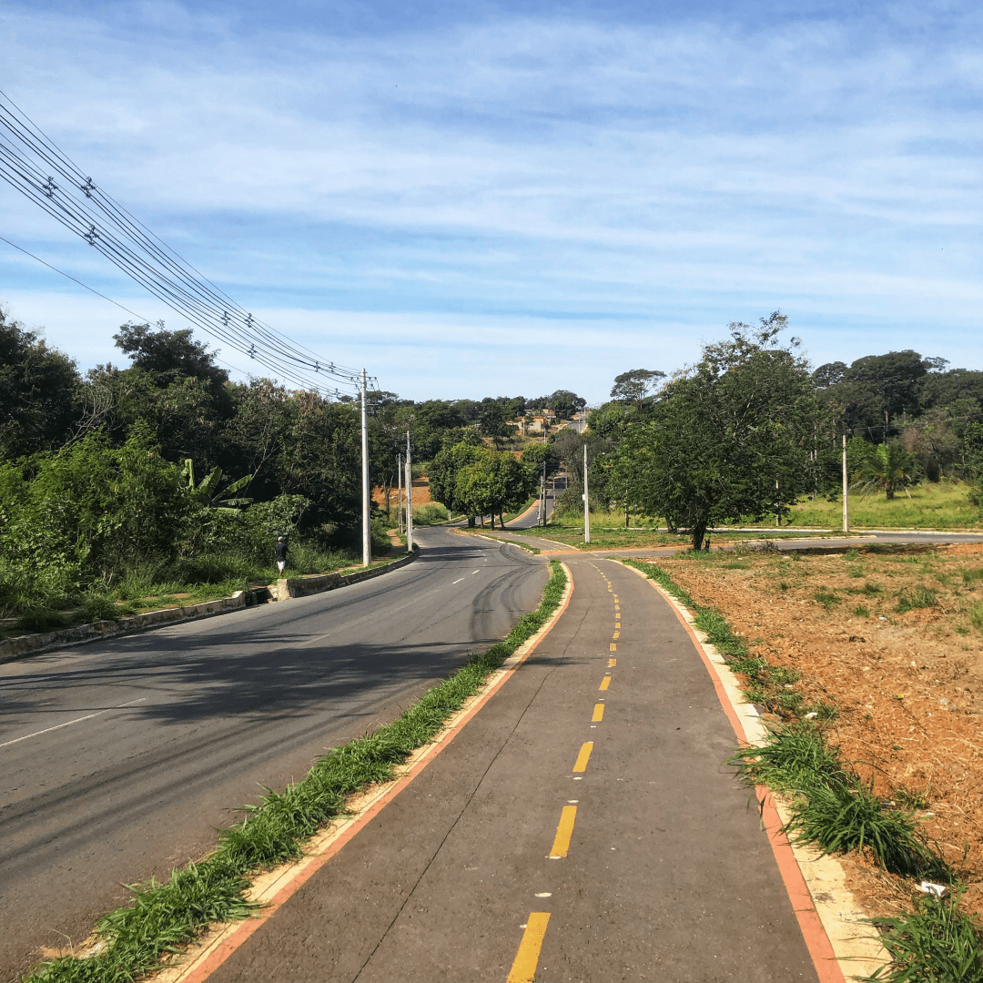 Fotografia de uma avenida de pista dupla, com uma ciclovia à esquerda, no canteiro central. A imagem foi capturada a partir da ciclovia, onde se pode observar a avenida descendo até um certo ponto, onde faz uma curva e sobe uma encosta íngreme. Tanto o canteiro central quanto as áreas ao redor da via estão repletos de árvores verdejantes. O céu está claro e intensamente azul, adornado por nuvens dispersas que se assemelham a fumaça.