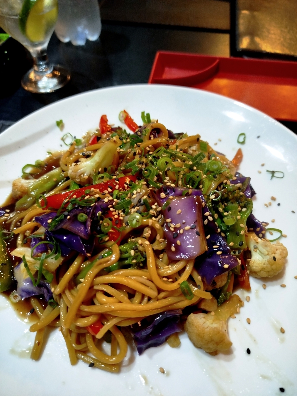 Foto de prato de Yakisoba de legumes. Macarrão e legumes sobre um prato branco, ao fundo mesa preta, prato quadrado vermelho e taça com água e limão.