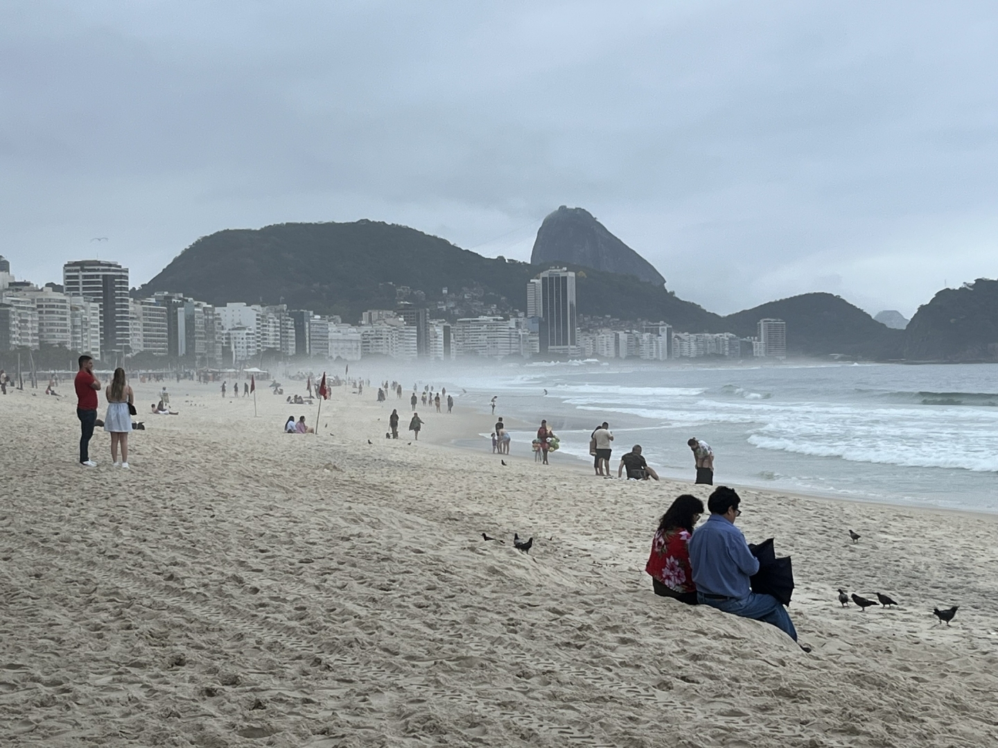 Fofo diurna na praia de Copacabana tirada ao nível dos olhos em um dia nublado, chuvoso e frio para os padrões cariocas. O céu acima coberto de nuvens acizentadas-azuladas, a faixa de areia forma um arco começando no lado direito da foto e seguindo pela esquerda até se perder na neblina no fundo da foto onde vemos os contornos dos morros no Leme. Logo abaixo deles, vindo do lado esquerdo da foro, uma faixa de prédios. À direita o mar também acinzentado-azulado praticamente sem ondas. A areia, que domina a maior parte da foto abriga pessoas esparsas observando o mar…