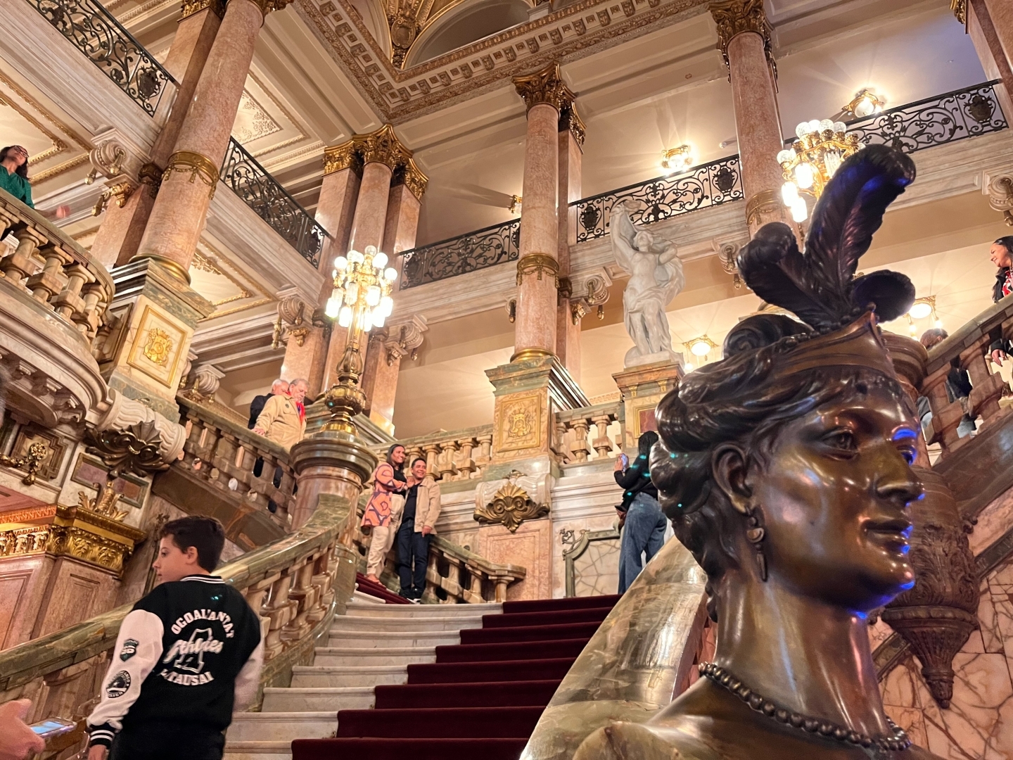 O enorme salão de entrada do Teato Municipal do Rio de Janeiro.
No centro da foto a escada, em primeiro plano à direita o busto de uma estátua de bronze com um adorno na cabeça.
Um jovem começa a subir as escadas do lado direito.
O espaço é muito iluminado em tons rosados de mármore.
Ao redor vê-se sacadas dos três andares superiores do teatro.