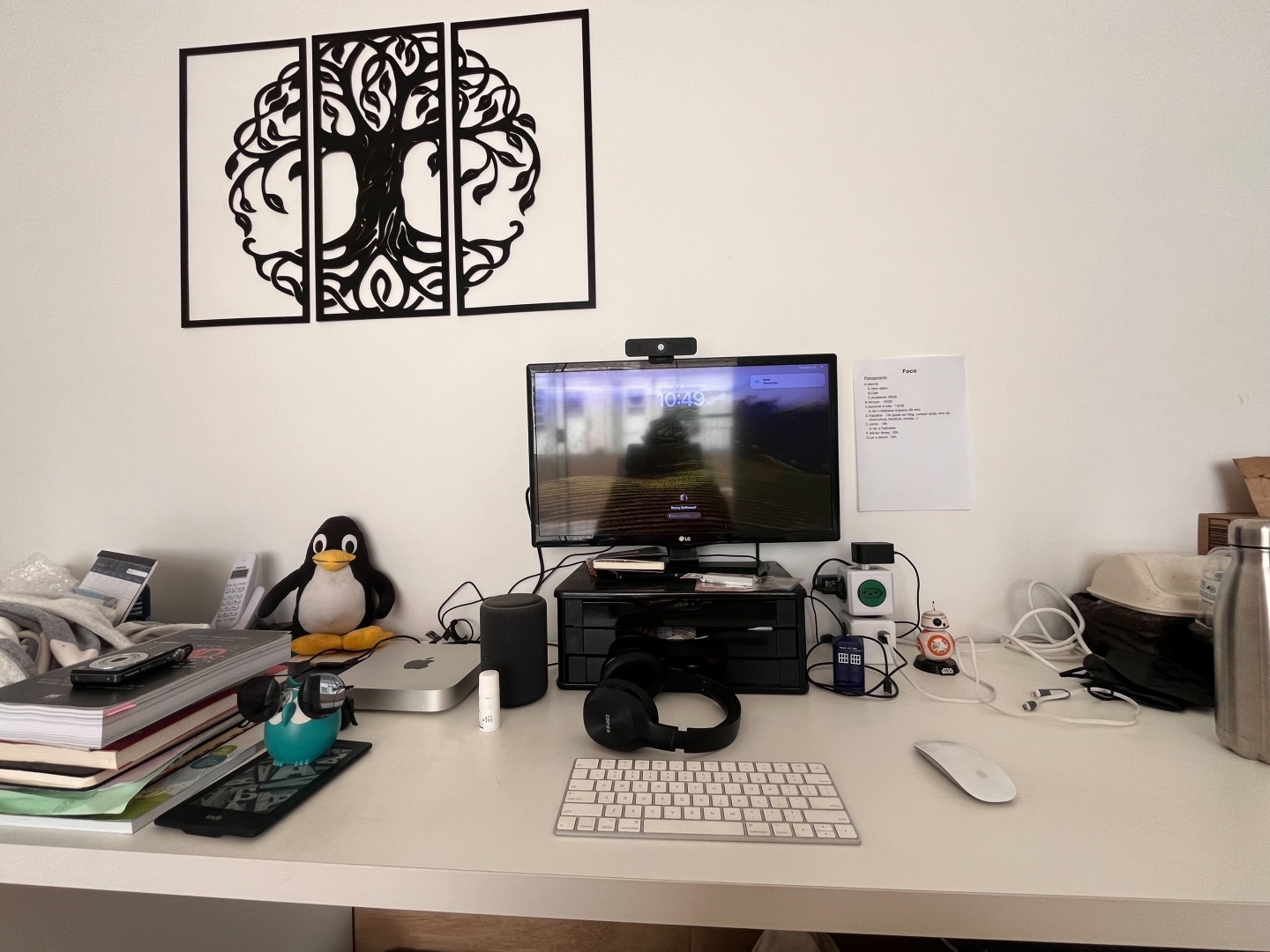 Mesa branca diante de uma parede também branca com uma árvore da vida preta. A maior parte da mesa está livre. No centro da mesa um teclado branco, um monitor sobre um suporte preto. Entre eles um headset. À direita um mouse também branco. Colado na parede um cubo com várias tomadas ligadas e alguns fios. À esquerda um Tux (pinguim do Linux) um Mac Mini e uma pequena bagunça de livros empilhados.