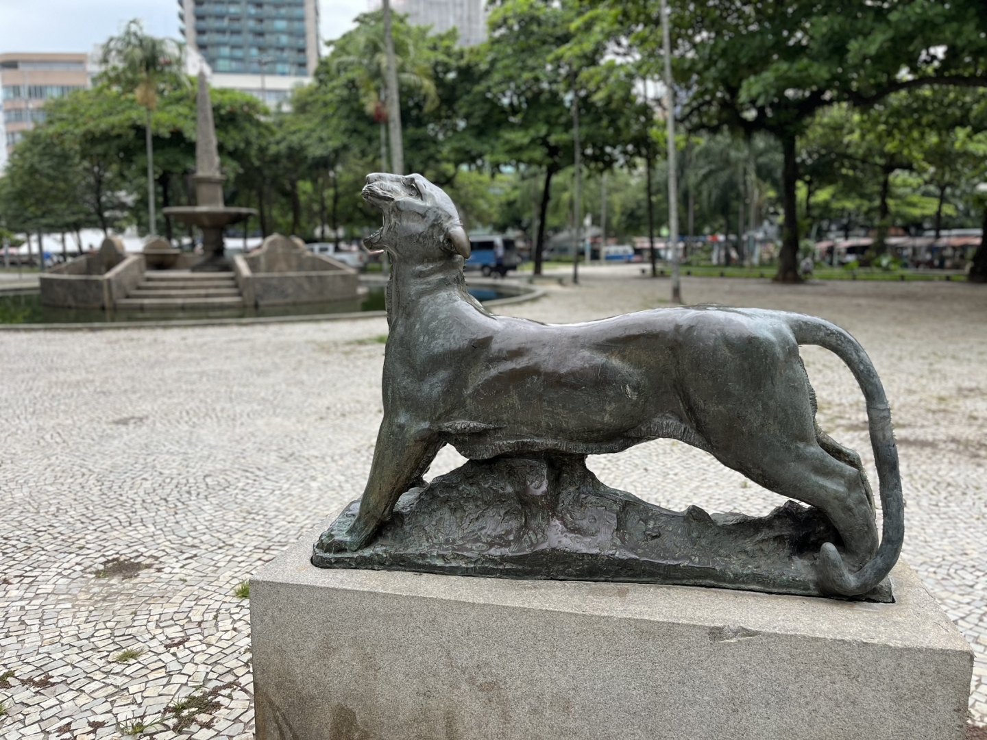 Foto de uma pantera de bronze em tamanho real vista de lado, sei corpo esticado e a cabeça levantada e rosnando para o alto.
Atrás vê-se uma praça de pedras portuguesas com árvores verdes ao fundo à direita e, à esquerda, um chafariz seco com um grande obelisco de pedra apontando para o céu. Mais além e também à esquerda, vemos alguns prédios.