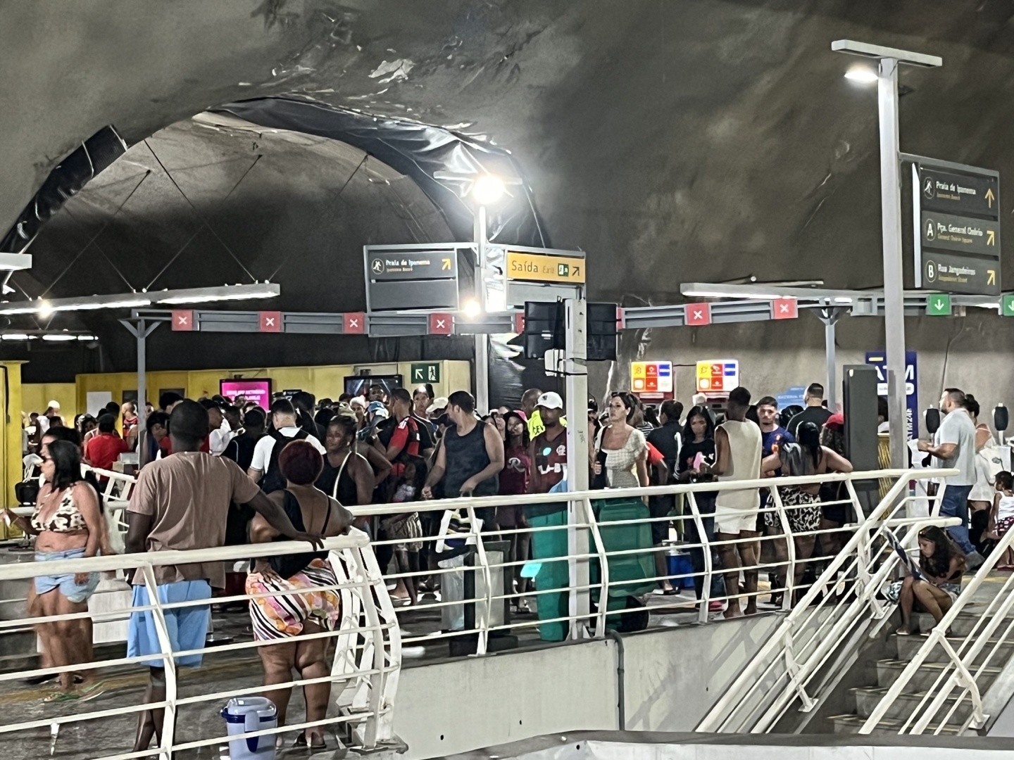 Foto de uma multidão de pessoas engarrafadas nas roletas do metrô. A grade branca em primeiro plano, que vai acima da cintura das pessoas, passa a impressão de um cercado para rebanhos.