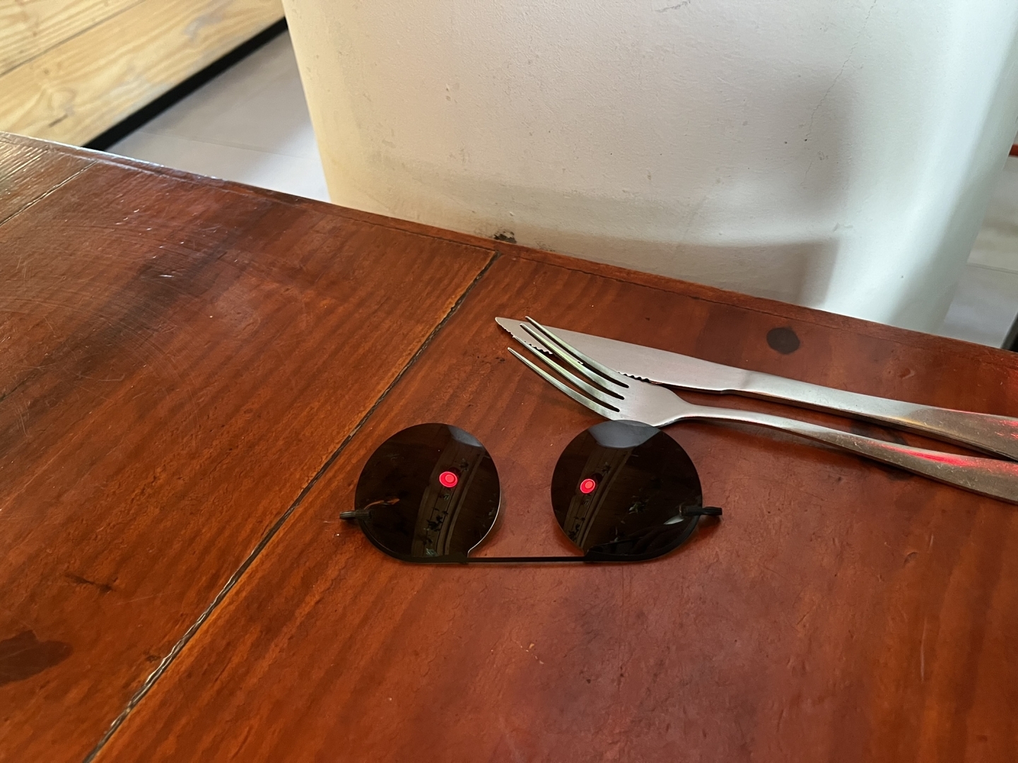 Um clip-on de lentes escuras refletindo uma luz vermelha em cada lente como se elas fossem olhos escuros com pupilas luminosas encarando a câmera com uma intensidade e seriedade quase humanas. As lentes estão sobre uma mesa de madeira envernizada e logo atrás delas um garfo e uma faca.