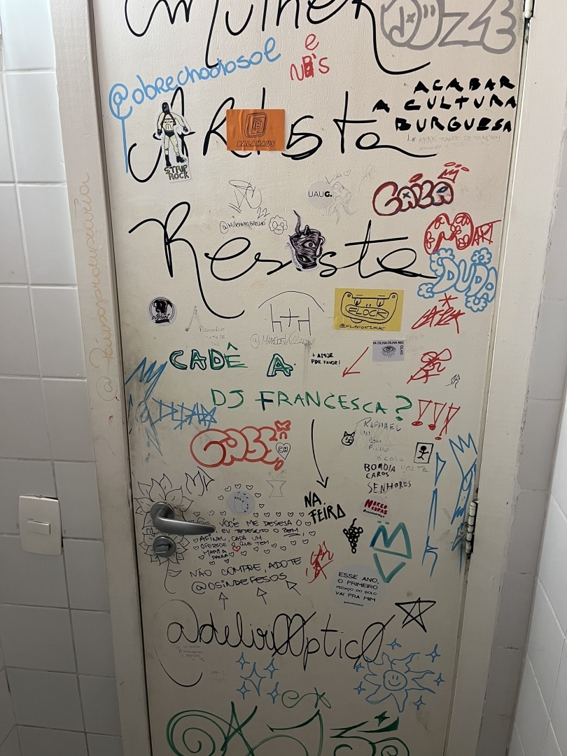Foto do lado interno da porta de um banheiro de restaurante.
A porta é branca e está coberta de grafites em várias cores: laranja, azul, verde, vermelho, mas predomina o preto.
Entre grafites com apelidos e mensagens como “onde está a dj Francesca” estão vários nomes de redes sociais, provavelmente Instagram. A maioria de perfil comercial.