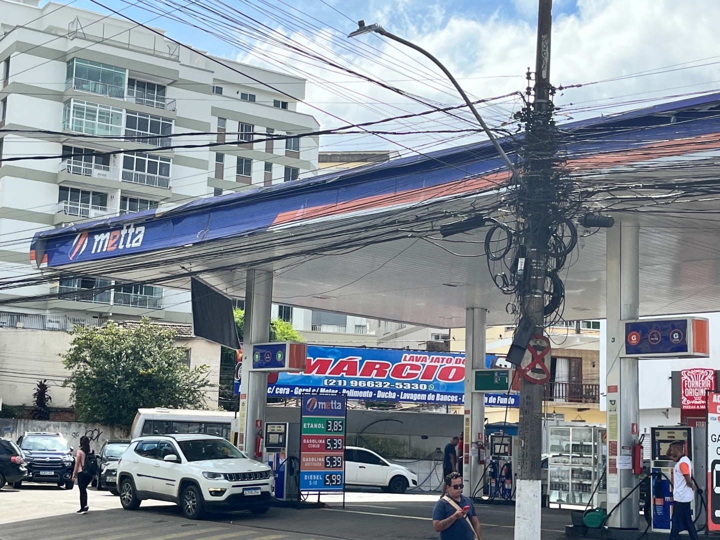 Posto de gasolina com uma cobertura de pilares altos com uma faixa azul onde se lê o nome do posto: Metta.
Em primeiro plano um poste de energia que sustenta uma infinidade de fios.
Um carro branco está parado no posto abastecendo. Ao fundo vê-se um prédio branco de sete andares. Três pedestres passam pelo posto