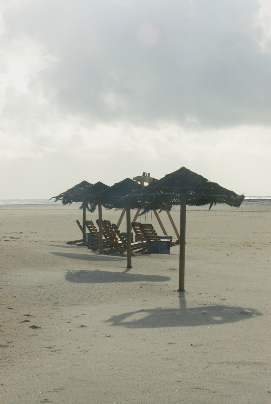 taken against the afternoon light
it is on a beach, in the middle of the picture are four thatched parasols.
there are wooden beach chairs and behind them the frame od a beach swing.
it appears to be a sepia-tone black and white, but is color.
across the middle of the shot is a thin line of the surf.
above are white and grey clouds.
the sand is a light sepia color