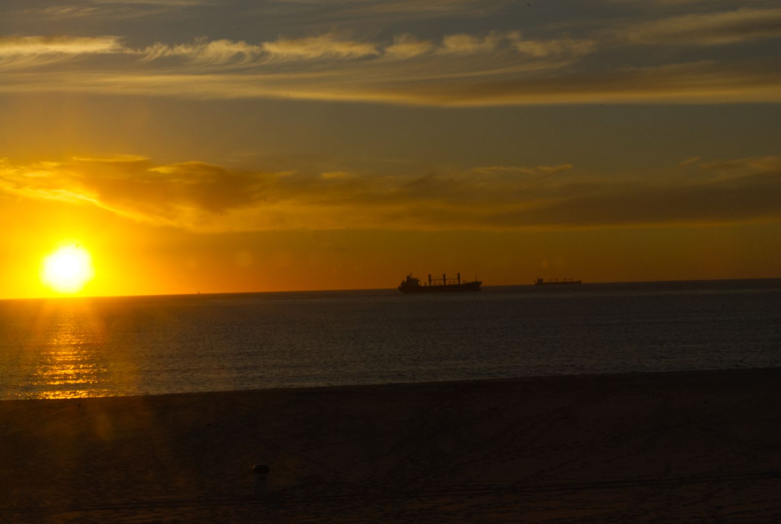 sunset picture broken up in bands running right to left
bottom is a band of extremely dark coffee, which is the sand.
the next band is the ocean, mostly calm.
the horizon line has two cargo ships on the right side.
the sun, not quite touching the horizon is on the left, ready to exit the stage.  
the sun is a bright yellow-orange fiery beach ball.
the sky, is a band of clouds, sky wispy clouds.