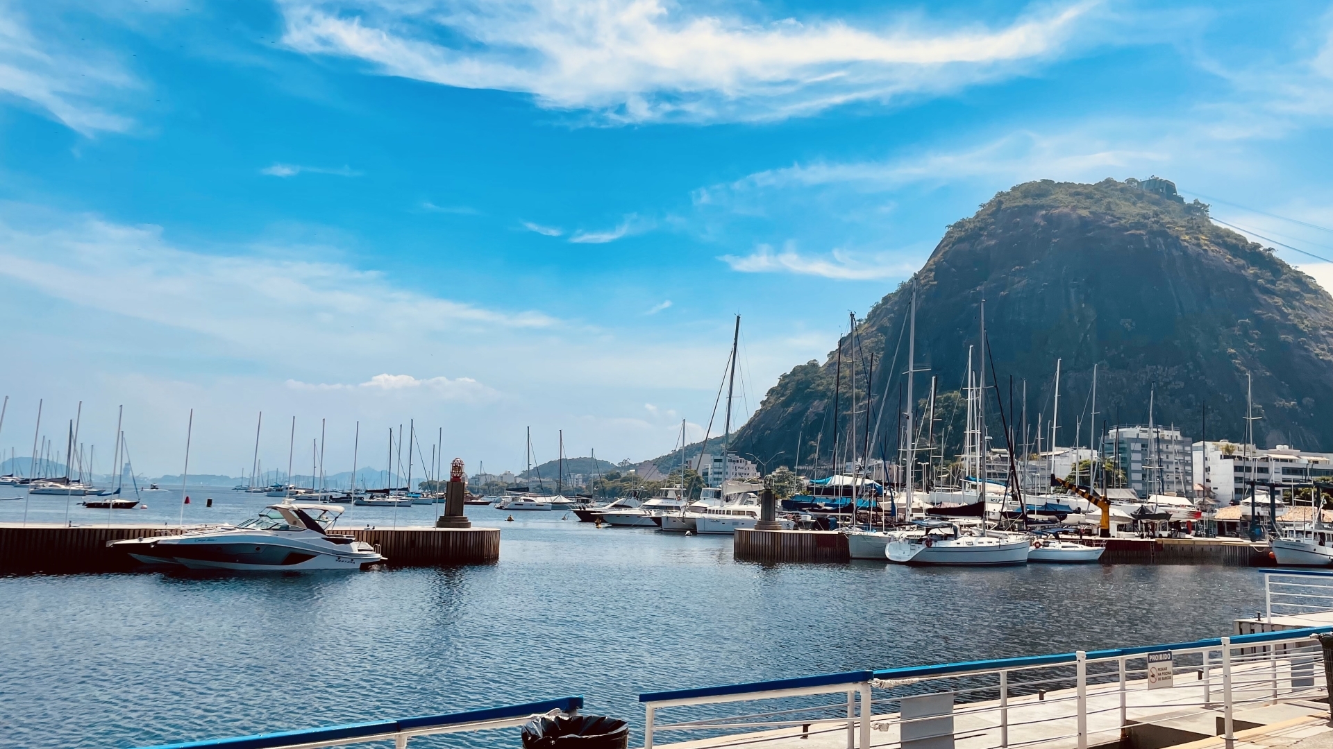 Imagem da vai de Guanabara com o Morro da Urca ao fundo. A imagem mostra uma marina com diversos barcos e veleiros ancorados em um píer. A atmosfera é tranquila, com um clima ensolarado.