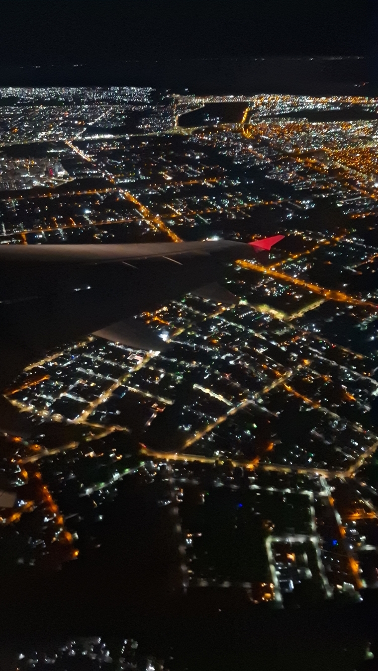 Uma foto tirada da janela de um avião mostrando o cenário do lado de fora, é possível ver a cidade de Brasília do alto a noite, com o céu escuro no horizonte e abaixo milhares de luzes da cidade, em contraste é possível ver assa do avião.