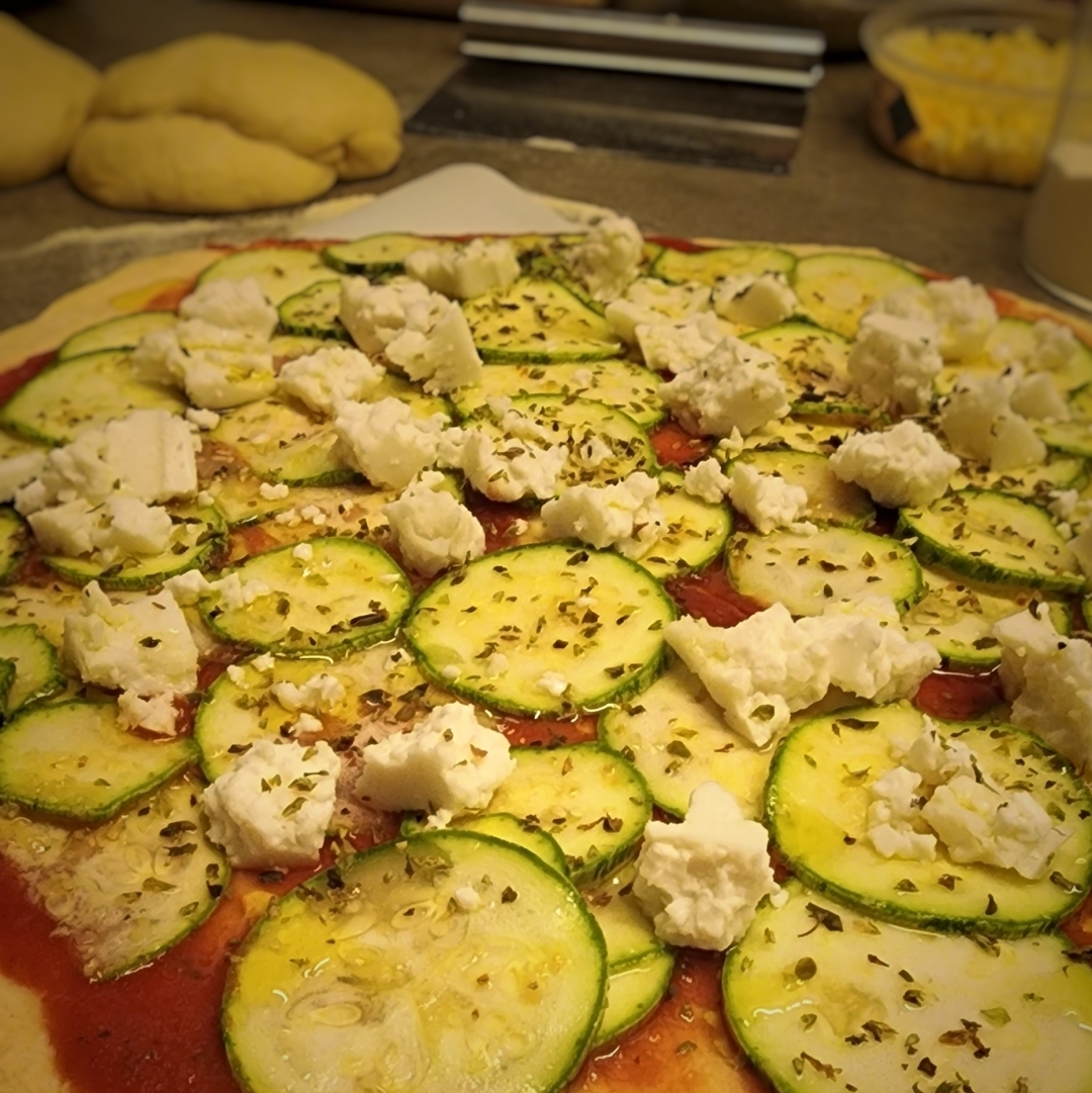 Foto em close de uma pizza antes de ir ao forno. a massa repousa sobre papel manteiga e tem sobre ela abobrinha, molho de tomate e queijo feita. ao fundo, é possível observar massas para outras pizzas, um cortador de massas e queijo ralado.