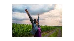 Woman with arms raised standing next to a cornfield under a cloudy sky, embarking on a journey within for self-discovery.