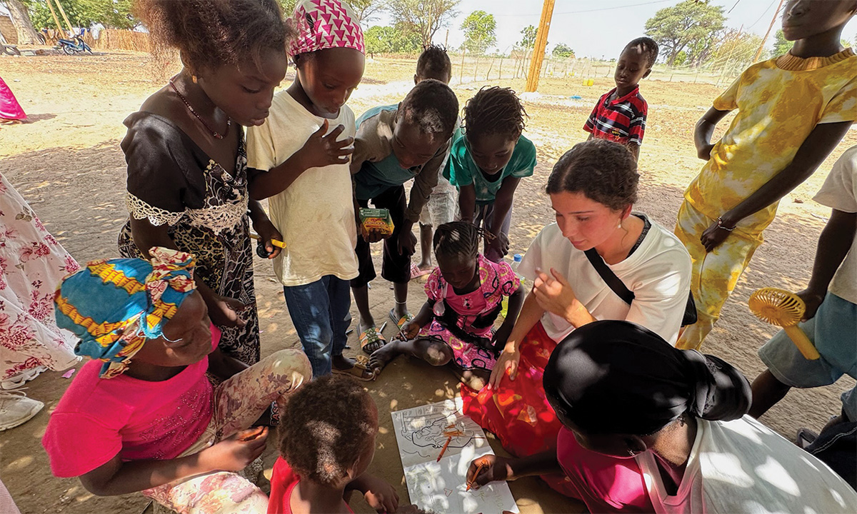 Mission trip volunteer sitting with a group of children outdoors, helping them draw and learn together under the shade of a tree.