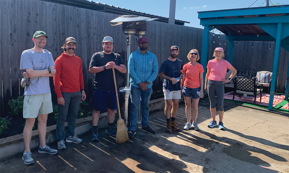 Group of volunteers standing outdoors with gardening tools in a backyard area during a service project.